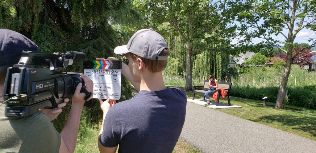 Two people from behind standing beside a path in a green park, one person is holding a camera filming the other person holding a clapperboard.