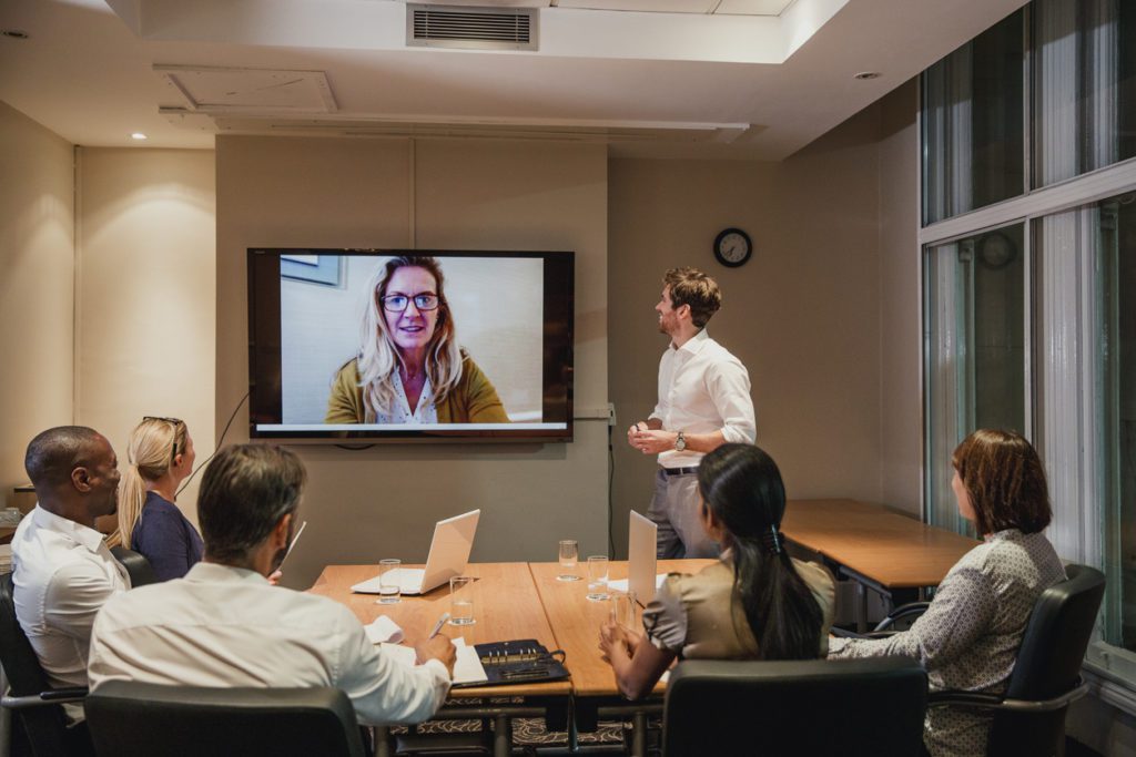 Group of people sitting at a desk in a small conference room, looking at one man standing and a woman on a TV, speaking through a video conference call.