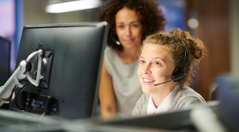 Two women looking at a computer screen. One woman is smiling and sitting closer to the screen wearing headphones and smiling. The other woman is standing behind her, slightly out of focus.